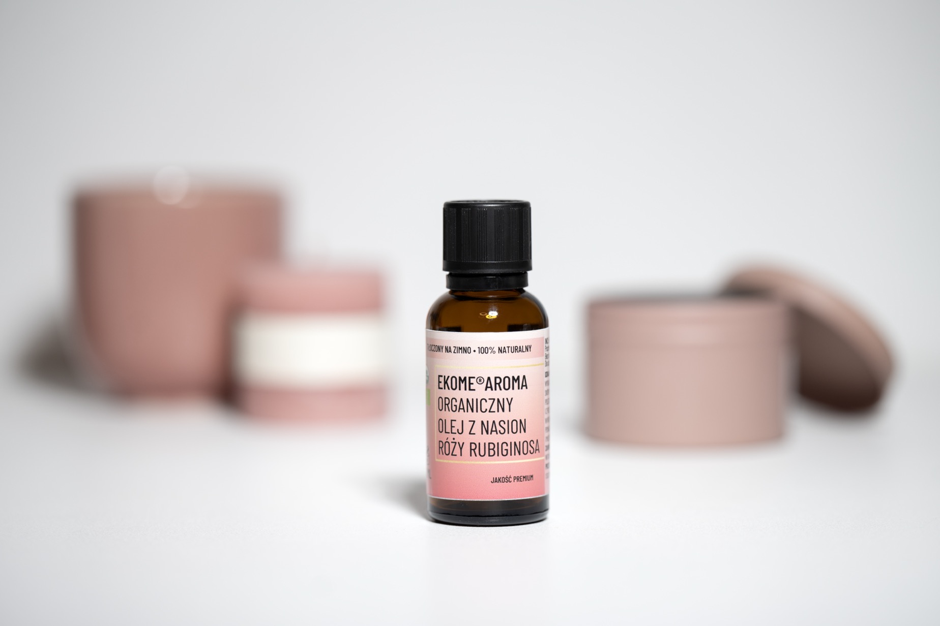 Dzika róża rubiginosa organiczny olej Ekome 30 ml w naturalnej aranżacji Brązowa butelka oleju z nasion dzikiej róży Rosa rubiginosa Ekome Aroma 30 ml na jasnym tle z różowymi akcesoriami
